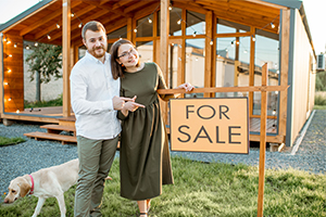 Smiling couple standing in front of a quaint country cottage with a 'For Sale' sign, surrounded by lush greenery and a serene atmosphere.