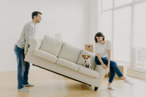 A couple carefully maneuvering a beige couch across the room while staging their house for sale.