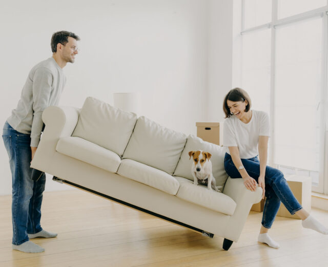 A couple carefully maneuvering a beige couch across the room while staging their house for sale.